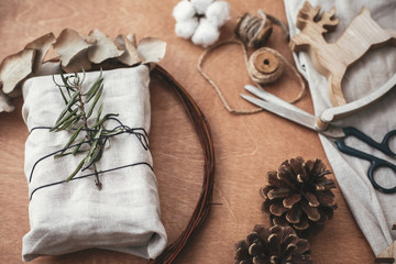 Stylish rustic gift wrapped in linen fabric with green branch on wooden table with pine cones,wreath,tree, reindeer, scissors, twine, cotton. Simple eco presents plastic free. Zero waste holidays