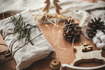 Stylish christmas rustic gift wrapped in linen fabric with green branch on wooden table with pine cones, reindeer, twine, lights. Simple eco presents plastic free. Zero waste  holidays.