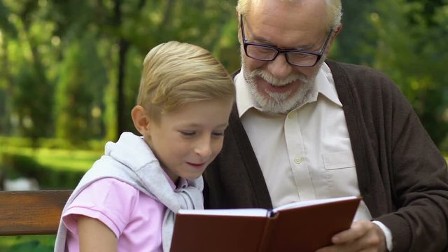 Little cute boy sitting on bench near grandfather showing him family photo album