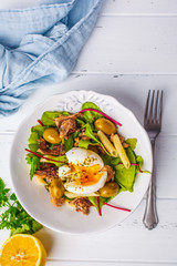 Tuna salad with pasta, olives and poached egg in white plate on white wooden background.