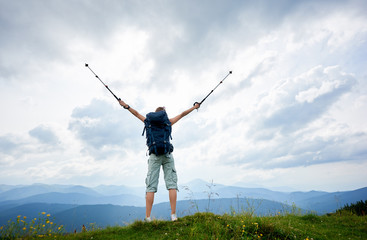 Back view of attractive woman hiker standing on a top of mountain with raised hands, holding trekking sticks, wearing backpack, enjoying summer day. Outdoor activity, lifestyle concept