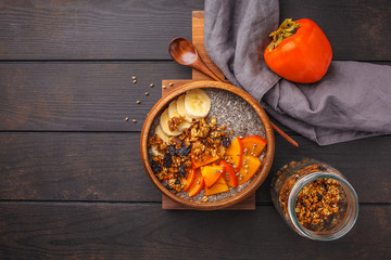 Chia pudding with banana, persimmon and granola, dark wooden background, top view, copy space.