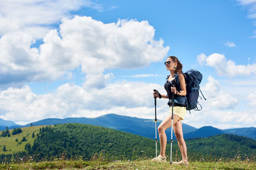 Naklejka premium Young woman backpacker hiking mountain trail, walking on grassy hill, wearing backpack and sunglasses, using trekking sticks, enjoying summer sunny day in the mountains. Tourism concept