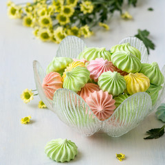 Sweet dessert. Multicolored air meringues in a round glass vase, next to a sprig of yellow chrysanthemums on a wooden table. Selective focus.