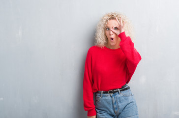 Young blonde woman with curly hair over grunge grey background doing ok gesture shocked with surprised face, eye looking through fingers. Unbelieving expression.