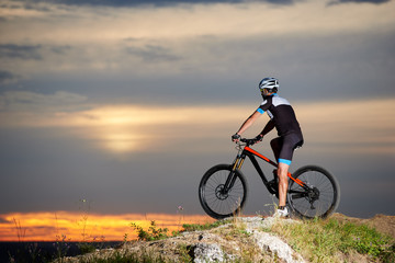 Side view of a biker on a mountain bike in sports clothes and a helmet on top of a rock resting on...
