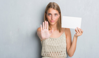 Beautiful young woman over grunge grey wall holding blank card with open hand doing stop sign with...