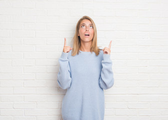 Beautiful young woman over white brick wall amazed and surprised looking up and pointing with fingers and raised arms.