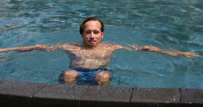 Happy man enjoying swimming pool vacation at tropical resort hotel. Male model at infinity pool relaxing in water on sunny summer day vacation getaway. 59.94 FPS