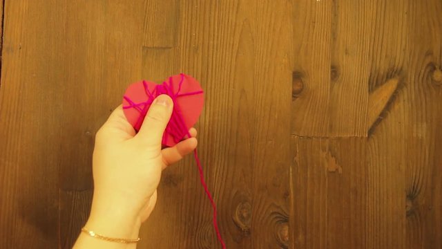 The girl makes a handicraft of red cardboard heart and pink thread. Time laps