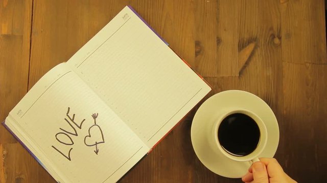 The girl writes in the diary the word love with a black felt-tip pen and drinks coffee. Time laps