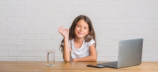 Young hispanic kid sitting on the table using computer laptop doing ok sign with fingers, excellent...