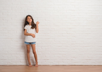 Young hispanic kid stading over white brick wall smiling with happy face looking and pointing to the side with thumb up.