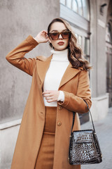 Outdoor portrait of young beautiful fashionable woman wearing trendy leopard print sunglasses, autumn coat, turtleneck, skirt, holding stylish handbag, posing in street of european city