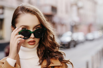 Outdoor close up portrait of young beautiful woman with long hair wearing big stylish green sunglasses, model posing in street of european city. Copy, empty space for text