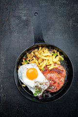 Traditional fried Allgauer veal roast roll sausage slices with fried egg in a cast-iron skillet with spices as closeup on a black board