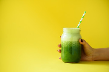 Woman hand holding glass jar of green smoothie, fresh juice against yellow background. Healthy...