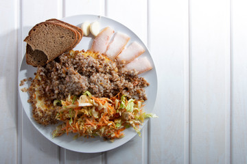 Buckwheat porridge, pieces of bacon, scrambled eggs, salad, and black bread in a white plate on a light wooden table