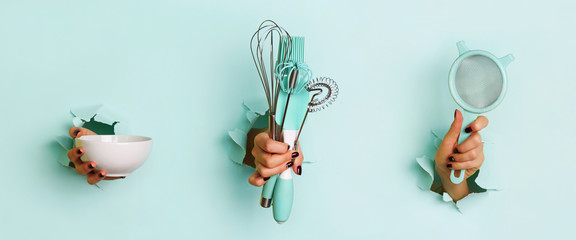 Woman hand holding kitchen utensils on blue background. Baking tools - bowl, sieve, brush, whisk,...