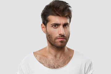 Serious young Caucasian man with dark thick stubble, trendy haircut, casual clothes, models against white background, frowns eyebrows, being angry with someone. Masculinity and lifestyle concept