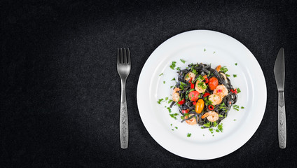 Black squid ink Fettuccine pasta with prawns or shrimps cherry tomatoes, parsley, chili in wine and butter sauce. Top view on granite with copy space.