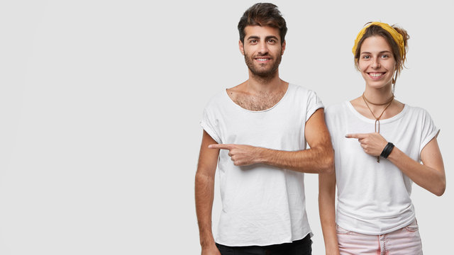 Photo of happy delighted woman and man dressed casualy, point at right side, show free space for your promotion or advertisement, pose in studio against white background, advertise new purchase