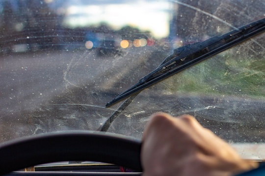 Dirty Scratched Car Windshield With Wiper Through Blurred Steering Wheel With Driver's Hand On Blurred Background