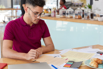 Naklejka premium Image of focused male student prepares report in finances, looks attentively at papers, eats delicious croissants, poses over cafe interior with free space for your promotion. Freelance work