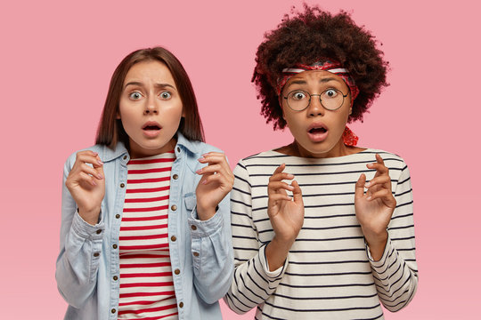 Photo of scared multiethnic women look with terror, notice something horrible, keep hands in protective gesture, wear striped clothes, isolated over pink background. People and fear concept.