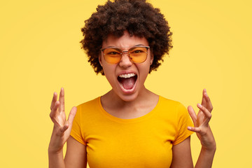 Its totally unfair! Displeased emotional mad dark skinned woman frowns and gestures angrily, expresses annoyance and anger, open mouth and gestures in madness, stands over yellow studio background