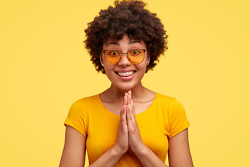 Studio shot of glad pleasant looking woman with dark skin, crisp hair, keeps both palms pressed together in praying gesture, wears bright casual t shirt, sunglasses, models against yellow background