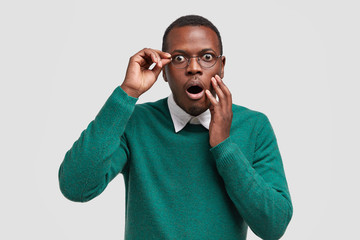 Scared dark skinned guy keeps jaw dropped from fear, notices horrible scene in front, dressed in green jumper, wears eyewear, expresses surprisement and fear, models over white background. Emotions