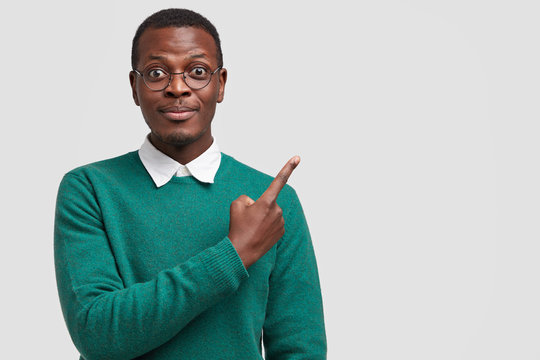 Pleased hipster male adult points with index finger at upper right corner, dressed in stylish clothes, demonstrates place where you can buy new outfit, isolated over white background with free space