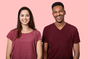 Fototapeta premium Joyful positive African American man and woman smile broadly at camera, have fun together, dressed in casual clothes, isolated over pink wall. Young mixed race couple stand indoor, giggles happily