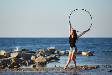 young girl in a black t-shirt and denim shorts dancing with a hoop on the beach © maksym
