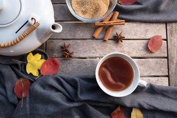 tea in a cup, a white teapot and spices on a wooden table with some autumn leaves, high angle view from above