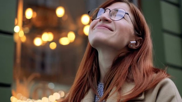 Beautiful Caucasian woman outdoor listening to music on smart phone. Young cute girl in headphones. Girl shaking her head to the rhythm of music with airpods outdoor