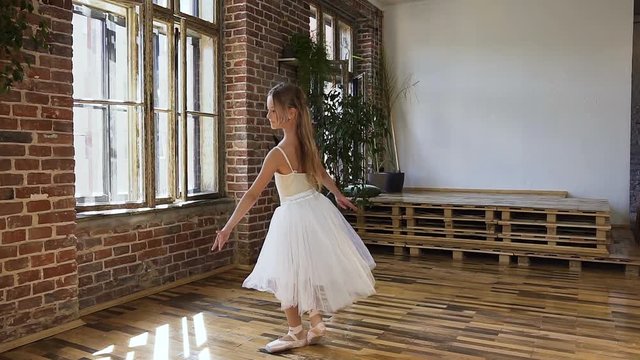 Young ballerina performs a classical ballet in pointe shoes and tutu on the background of a large window and red brick wall. Elegant ballerina dancing in pointe shoes and in a white fluttering dress