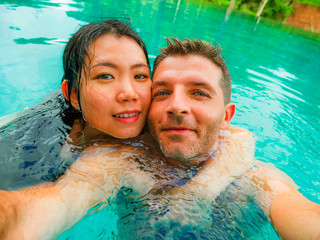 young happy and beautiful mixed ethnicity couple Asian woman and Caucasian man taking romantic selfie picture at tropical resort swimming pool enjoying honeymoon trip