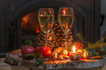 Christmas and New Year holiday concept: two glasses of champagne, fruits, nuts, burning candle on the tree stump in front of the fireplace