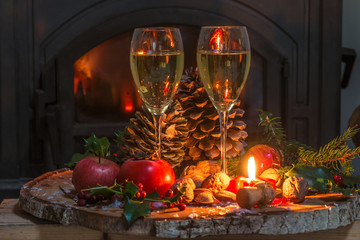 Obraz na płótnie Canvas Christmas and New Year holiday concept: two glasses of champagne, fruits, nuts, burning candle on the tree stump in front of the fireplace