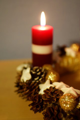 Red first advent candle burning with traditional christmas decoration wreath on wooden table at shallow depth of field with focus on pine cone. Xmas candlelight in dark.