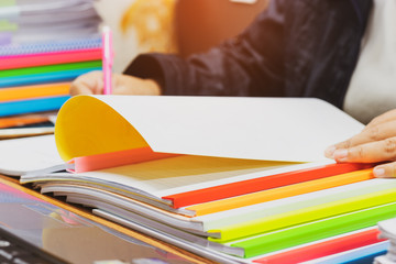 Teacher hand is holding pen for checking student homework assignments on desk in school. Unfinished paperwork stacked in archive with color plastic slide binder bars. Education and business concept.