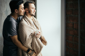 Nice same-sex married couple is standing together at window. Careful and gentle male brunette embracing from the back his woman-like male friend wrapped in soft blanket. They are happy.