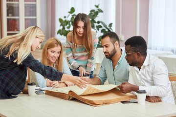 Multiethnic cheerful young people sitting at table reading big old reference book for preparing scientific report. Group of young students doing together homework at library.