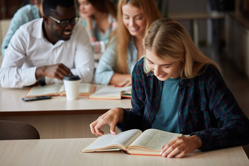 Smiling attractive blonde girl makes some notes while reading book in college library, looking at camera, sitting at desk with the other students on background