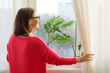 Woman looking out the window opening the curtains, view from the back