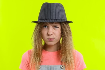 Portrait of a pretty funny teen girl with a pensive face, blonde girl with curly hair in hat with thoughtful expression, presses lips together, yellow studio background