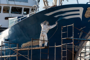 pintor pintando el casco de un barco , el pintor está protegido con una máscara y un buzo blanco y está trabajando sobre un andamio 