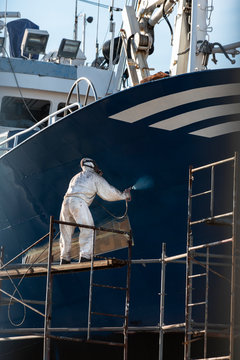 pintor pintando el casco de un barco , el pintor est&aacute; protegido con una m&aacute;scara y un buzo blanco y est&aacute; trabajando sobre un andamio 